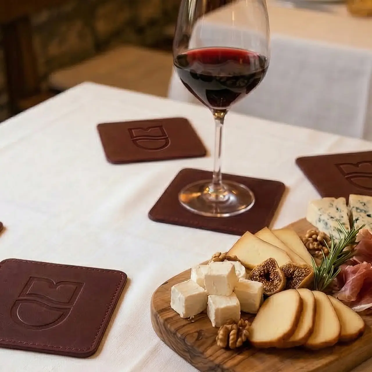 Wine glass on a brown coaster next to a wooden board with cheese, figs, walnuts, and prosciutto.