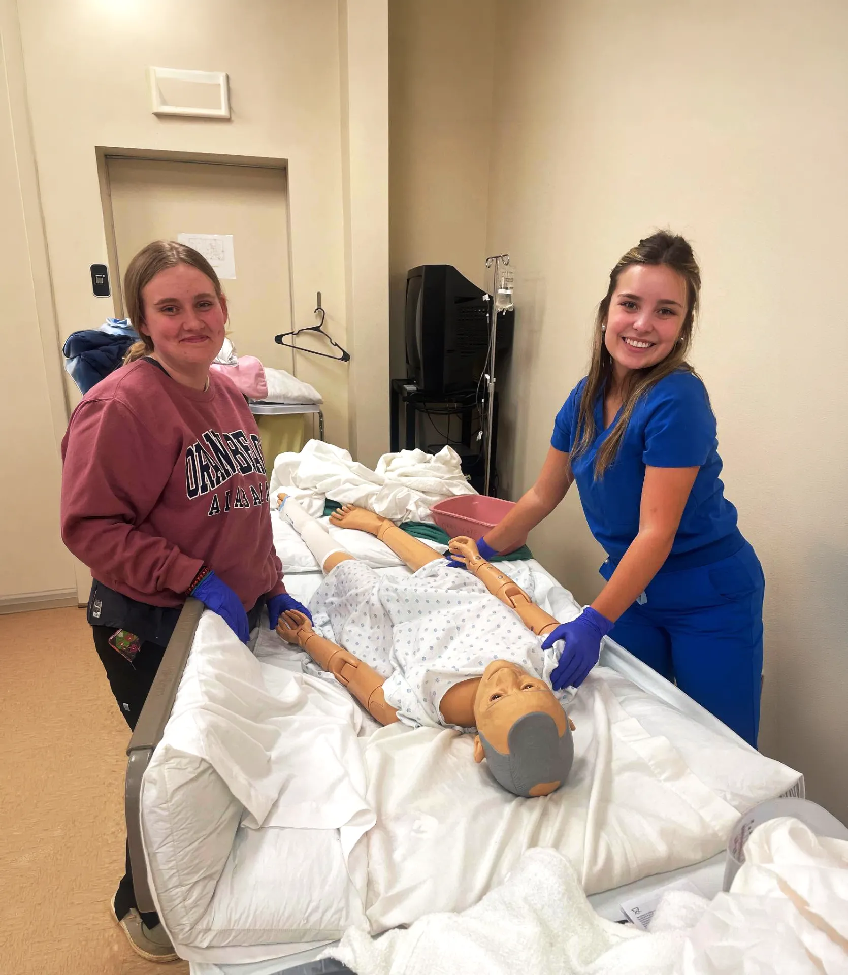 Two Compass Career College Practical Nurse women students practice patient care on a hospital bed mannequin in a medical training room.