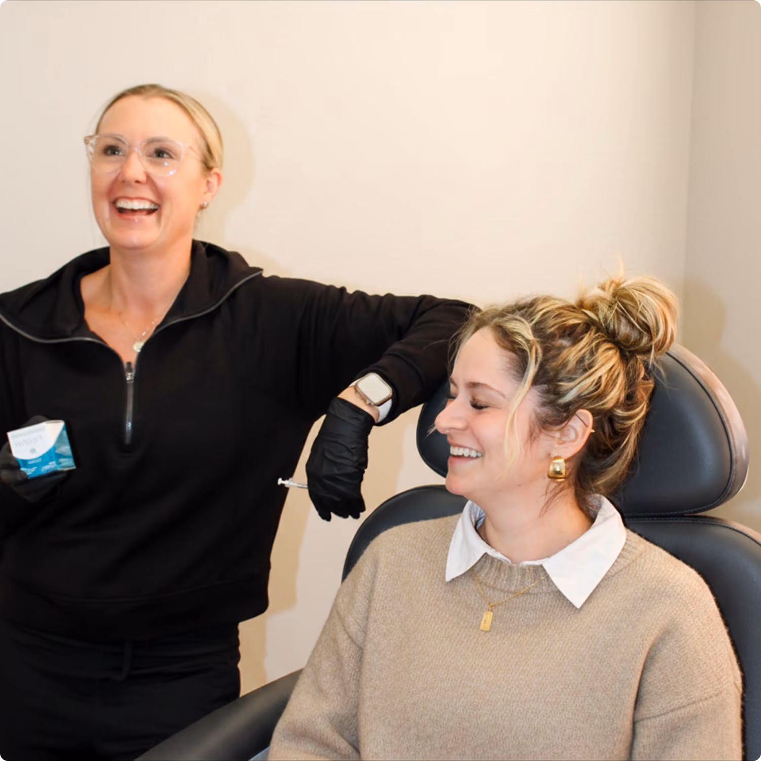 Smiling healthcare worker with gloves holding a syringe near a seated woman with blonde hair in a bun.