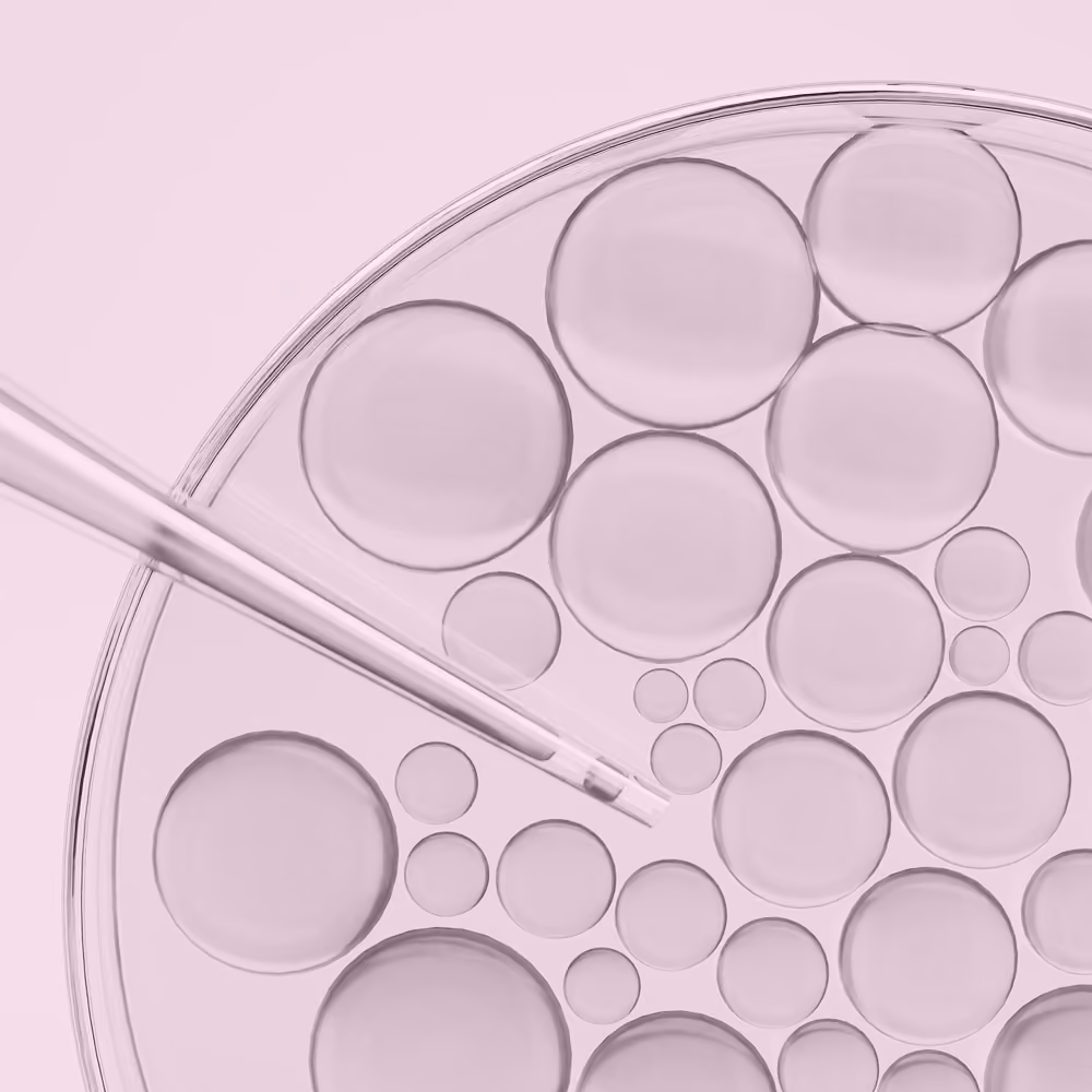 Glass pipette over a petri dish with various transparent liquid droplets on a pale pink background.