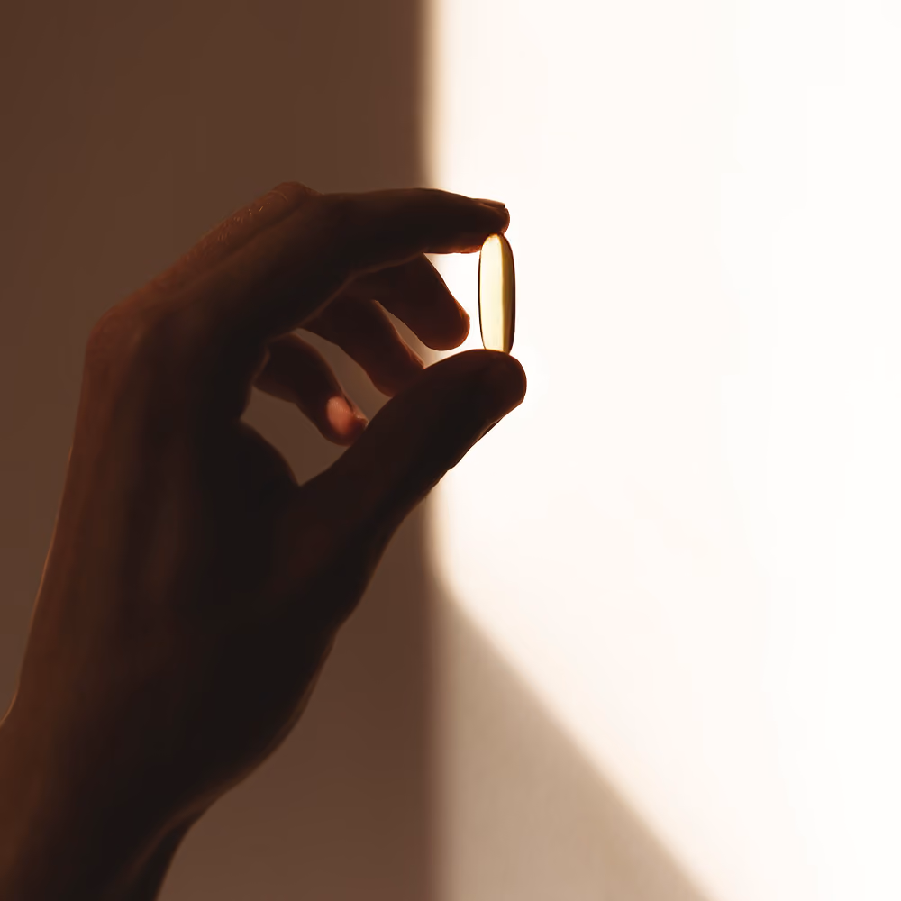 Hand holding a translucent yellow capsule pill against a bright background.