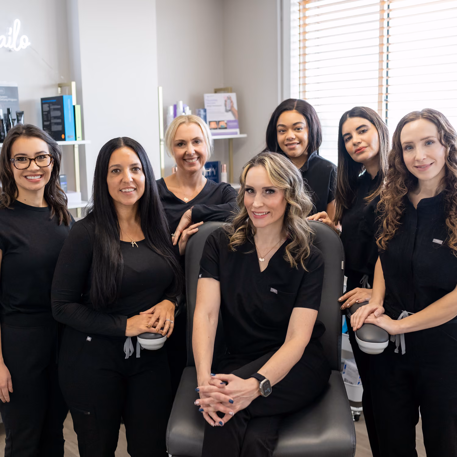 Smiling team of seven women in black scrubs posing in a bright, modern medical or spa office.