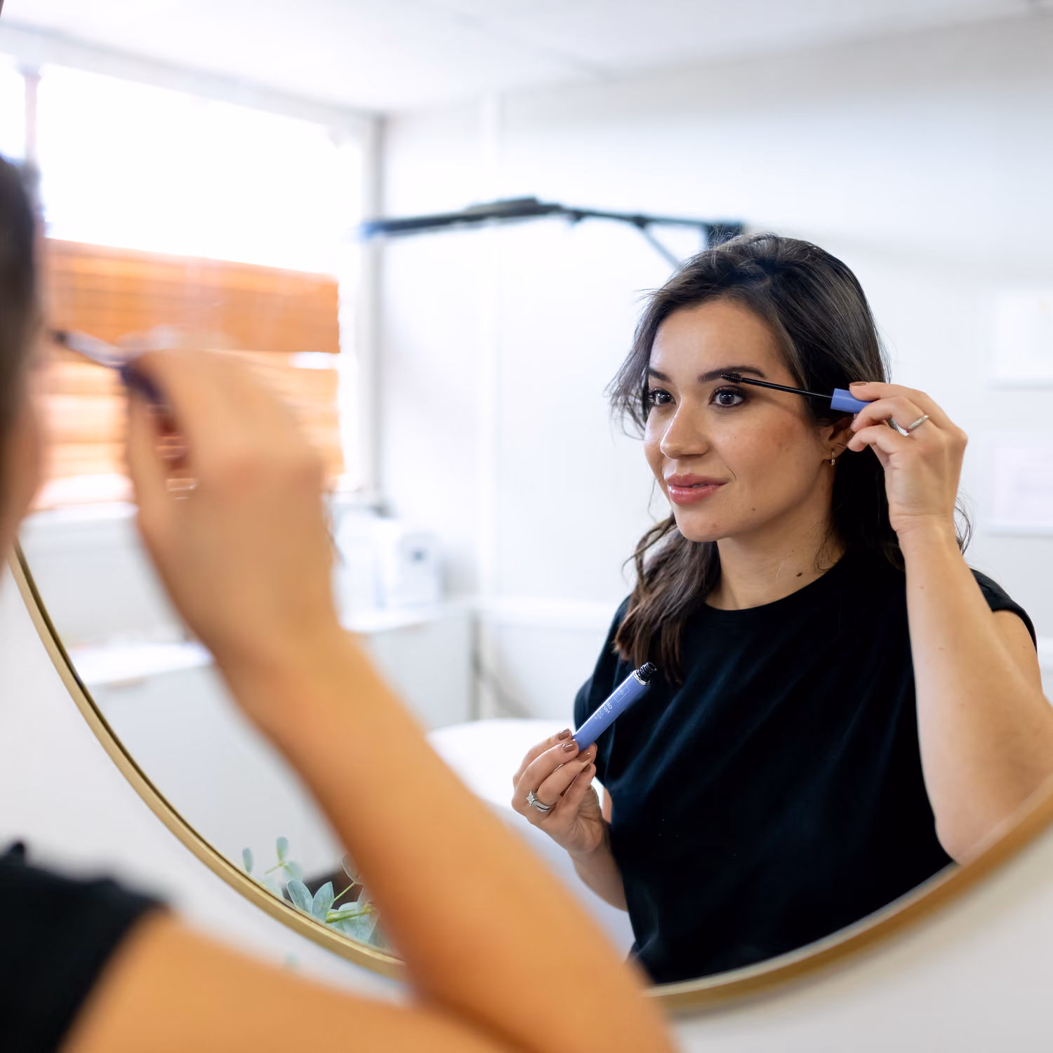 Woman applying mascara to her eyelashes while looking in a round bathroom mirror.