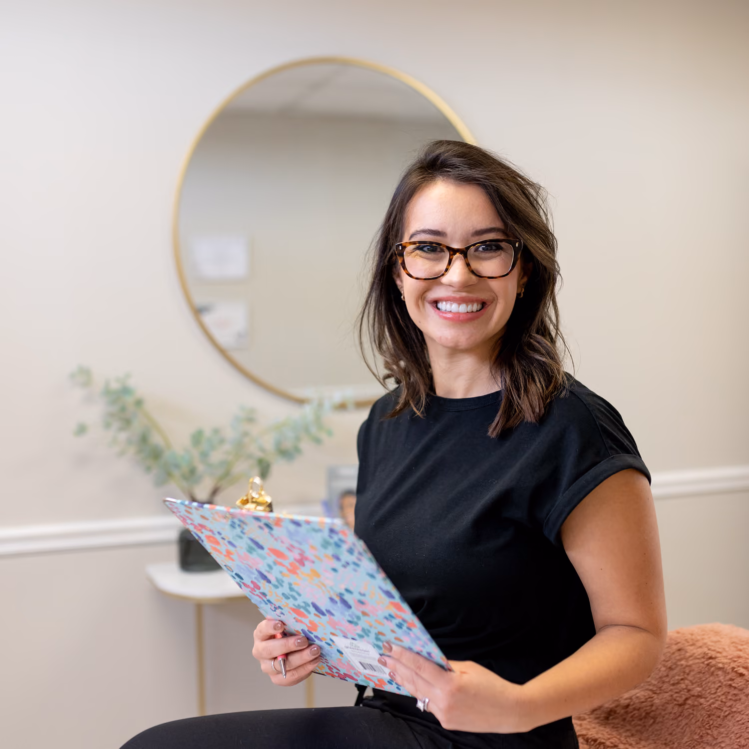 Smiling woman in glasses holding a colorful clipboard sitting in a bright room with a round mirror behind.