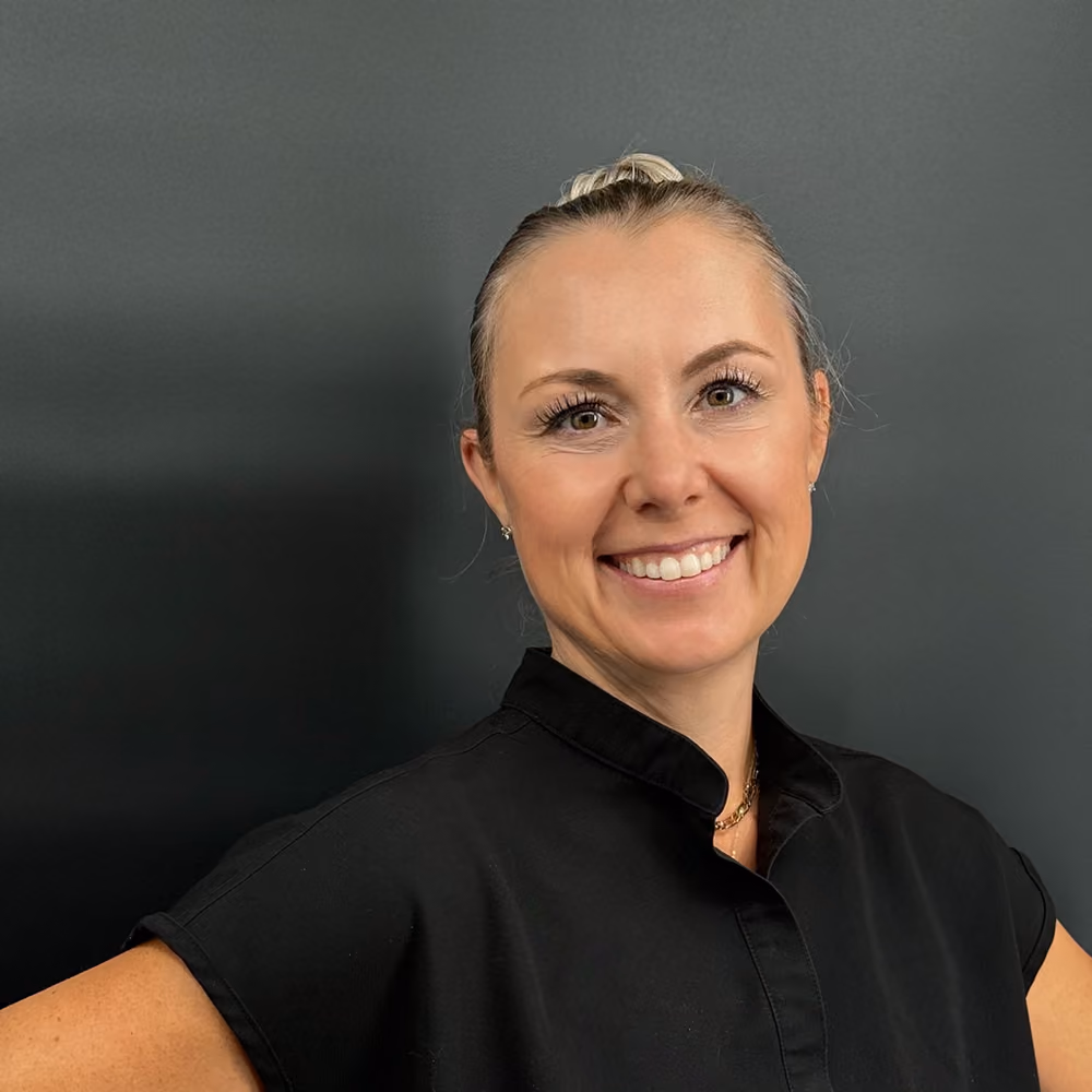 Smiling woman with blonde hair tied back wearing a black shirt against a dark gray background.