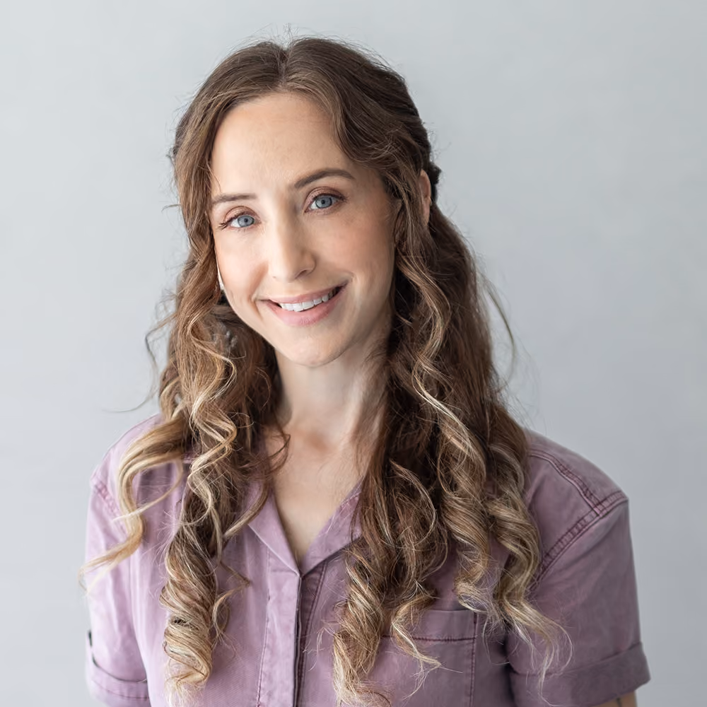 Smiling woman with curly brown hair wearing a light purple shirt against a plain gray background.