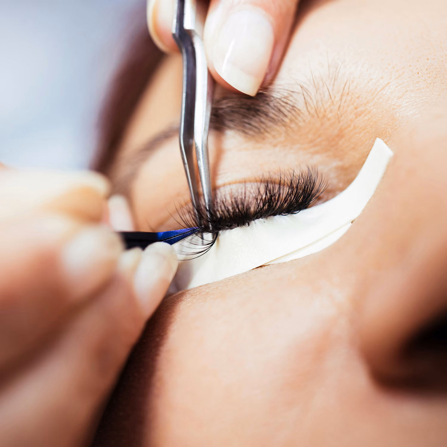 Close-up of eyelash extension application using tweezers on closed eye with protective pad.