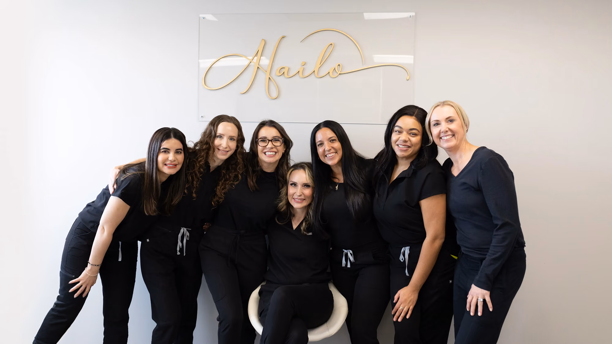 Seven women dressed in black uniforms smiling in front of a wall sign reading 'Hailo'.