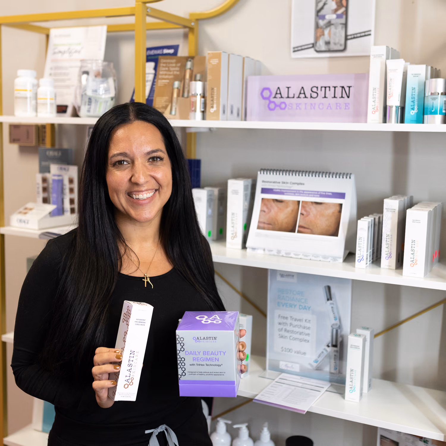 Smiling woman holding Alastin Skincare products in a store with shelves of skincare items behind her.
