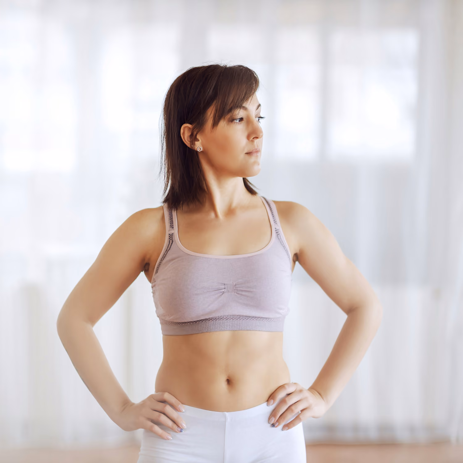 Fit woman in sports bra and leggings standing with hands on hips looking to the side in bright room.