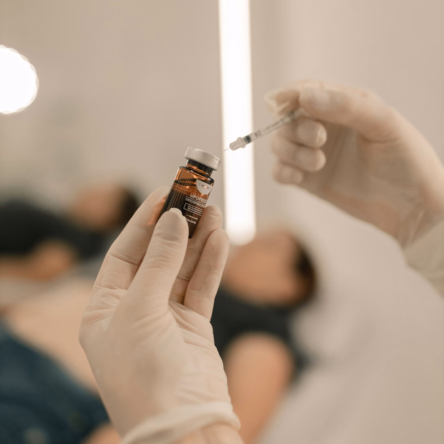 Hands in gloves holding a vial and syringe, with a person lying down blurred in the background.