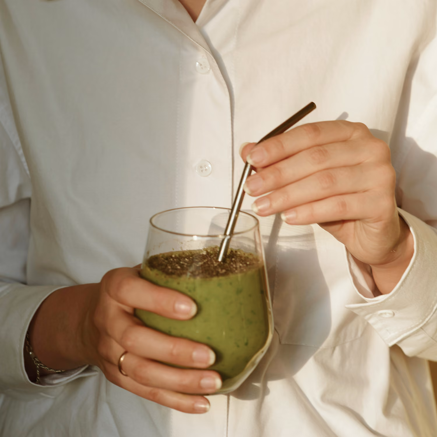 Person in white shirt holding a glass of green smoothie stirred with a metal straw.