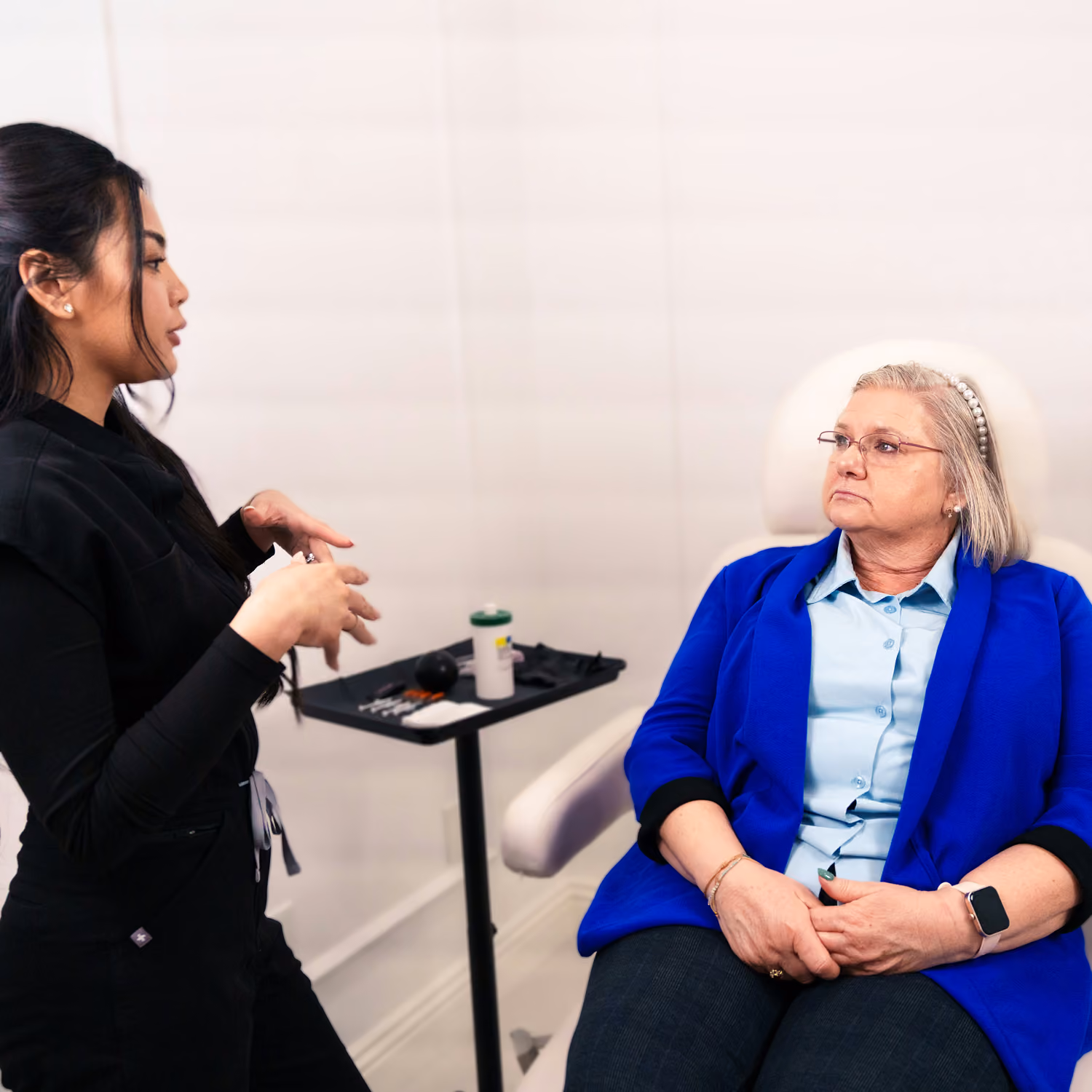 Woman in black discusses with seated older woman in blue blazer in a clinical setting.