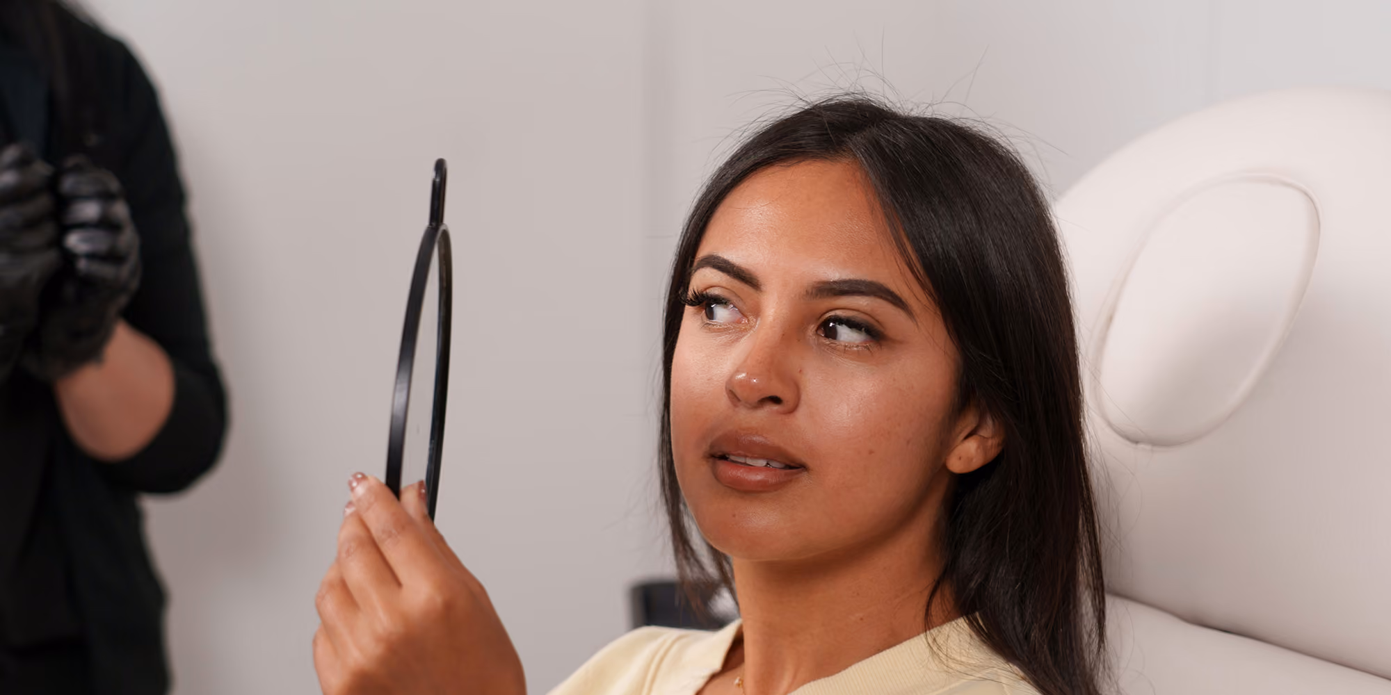 Woman with dark hair holding a black handheld mirror looking to the side in a clinical setting.