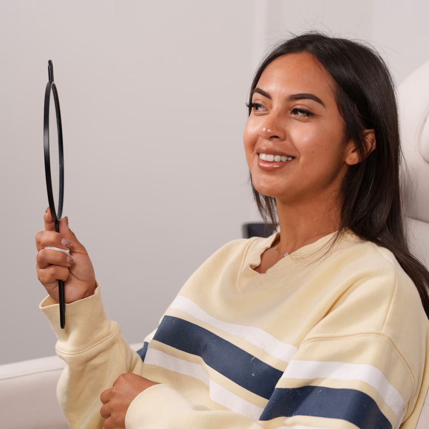 Smiling woman sitting and holding a round mirror with black handle, wearing a striped sweater.
