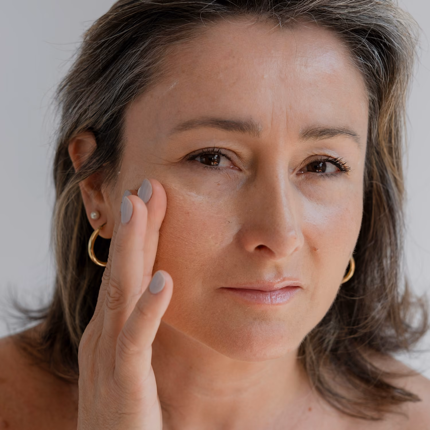 Woman with light makeup gently touching her cheek and looking at the camera.