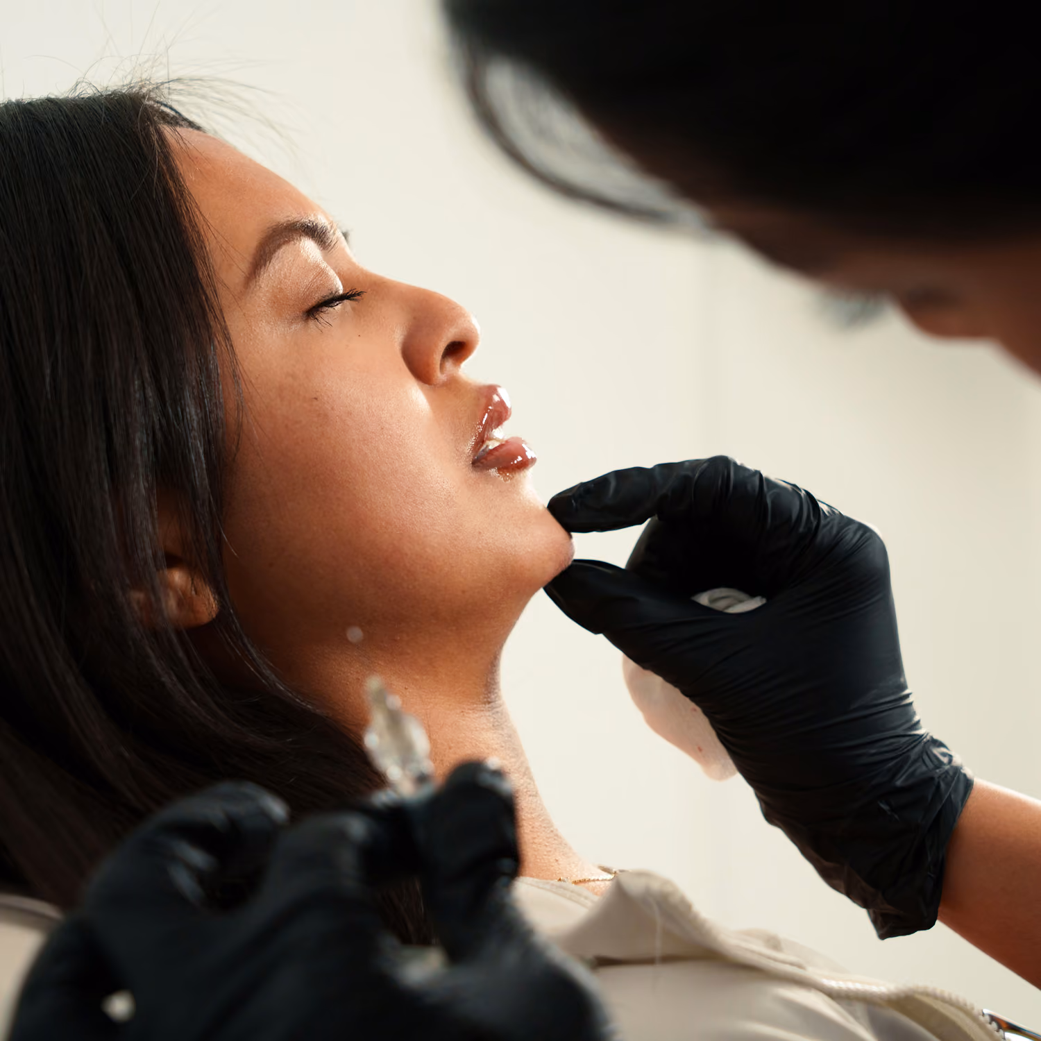 Woman with closed eyes receiving a cosmetic injection in her chin from a gloved professional.