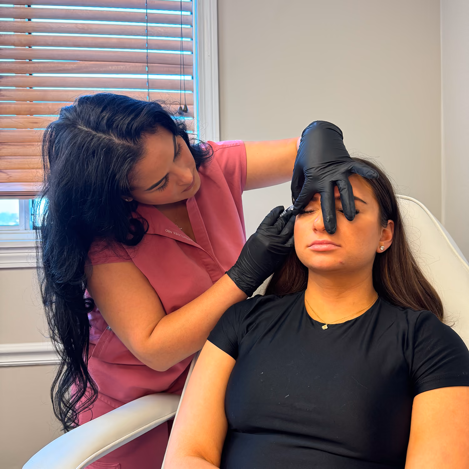 A woman in medical gloves examines another woman's face near her eye while she sits in a chair with eyes closed.