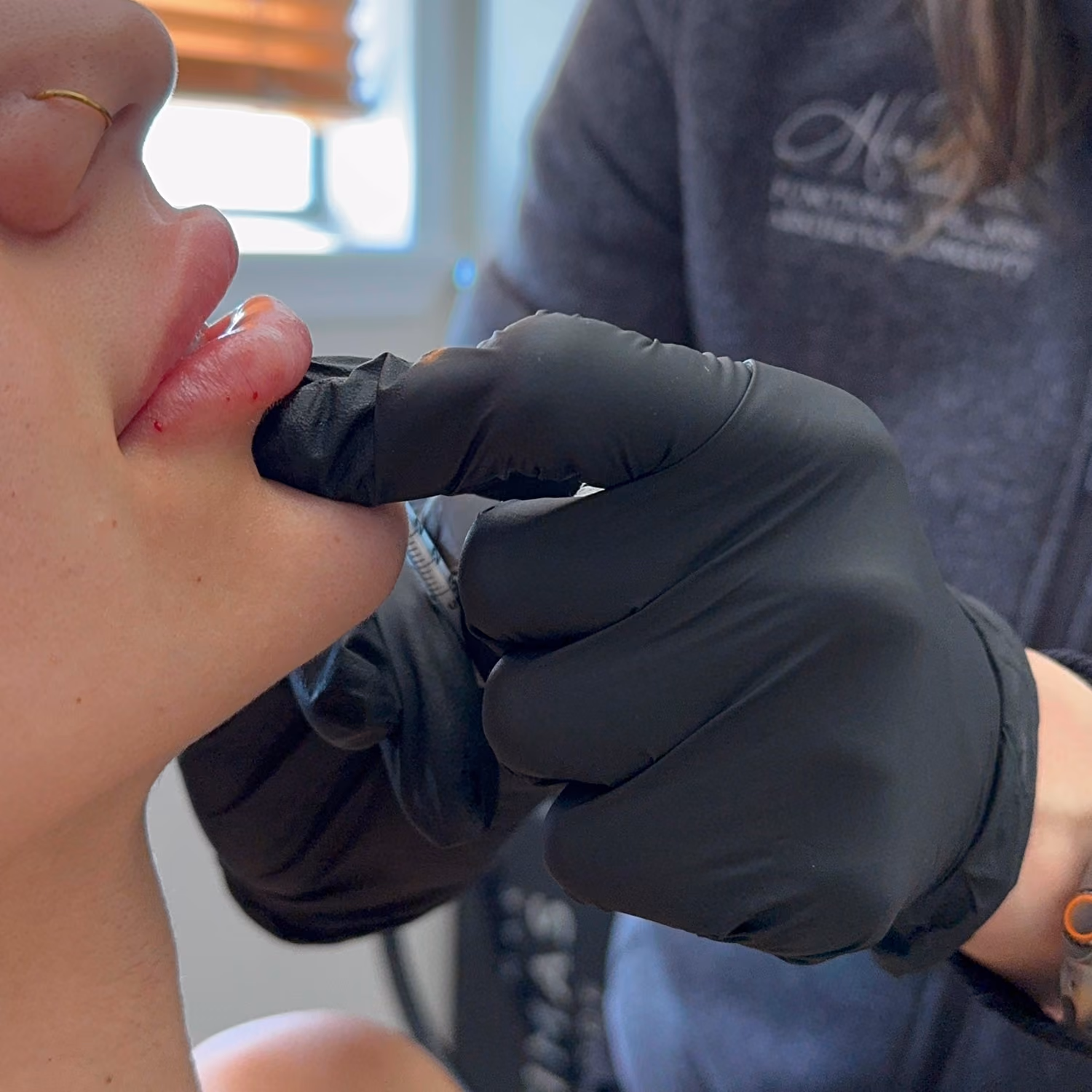 Person wearing black gloves preparing to pierce or tattoo the lower lip of another person.