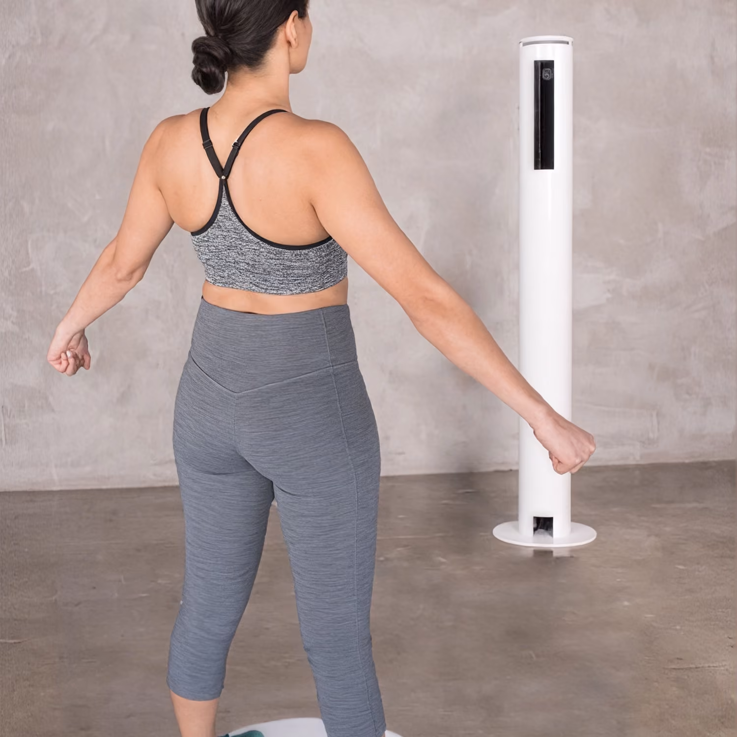 Woman in gray activewear standing on a circular platform facing a tall white scanning device.