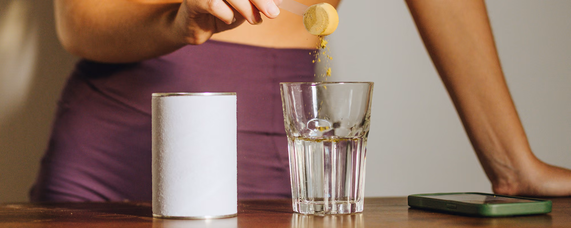 Person in purple pants pouring yellow powder from a scoop into a clear glass of water on a wooden table.