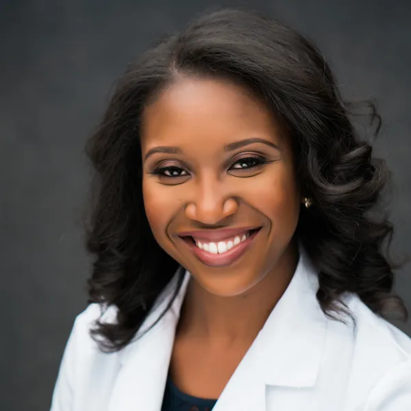 Portrait of a smiling woman with curly hair wearing a white coat and dark top against a dark background.