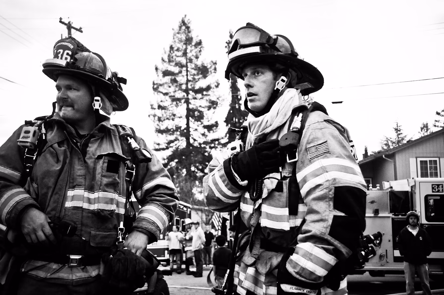 Two firefighters in full gear standing outside with fire trucks and people in the background.