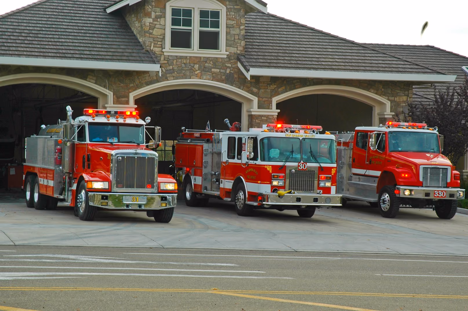 Three red fire trucks parked outside a stone fire station with garage doors open.