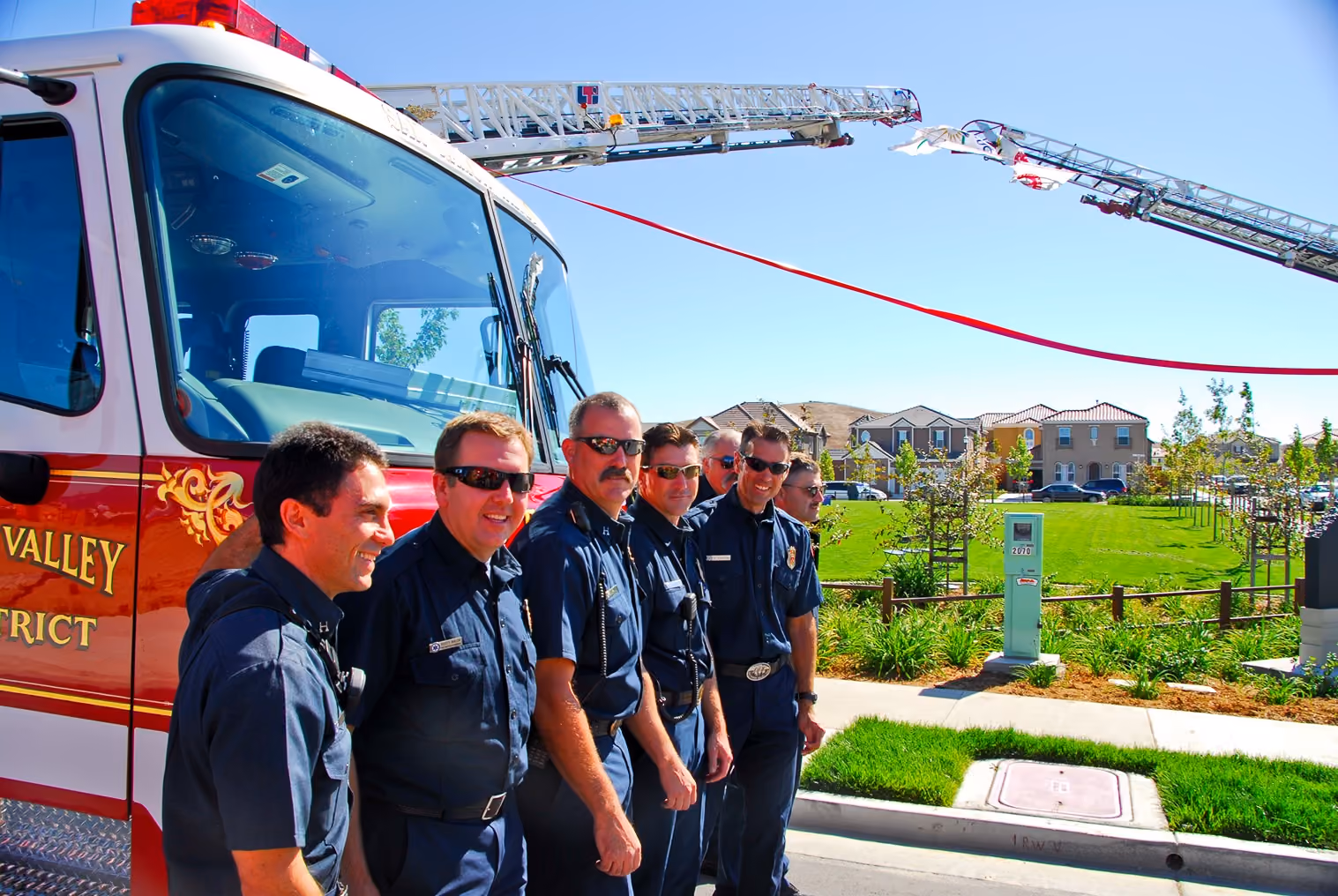 Group of six firefighters standing in front of a fire truck with extended ladders under a clear blue sky.
