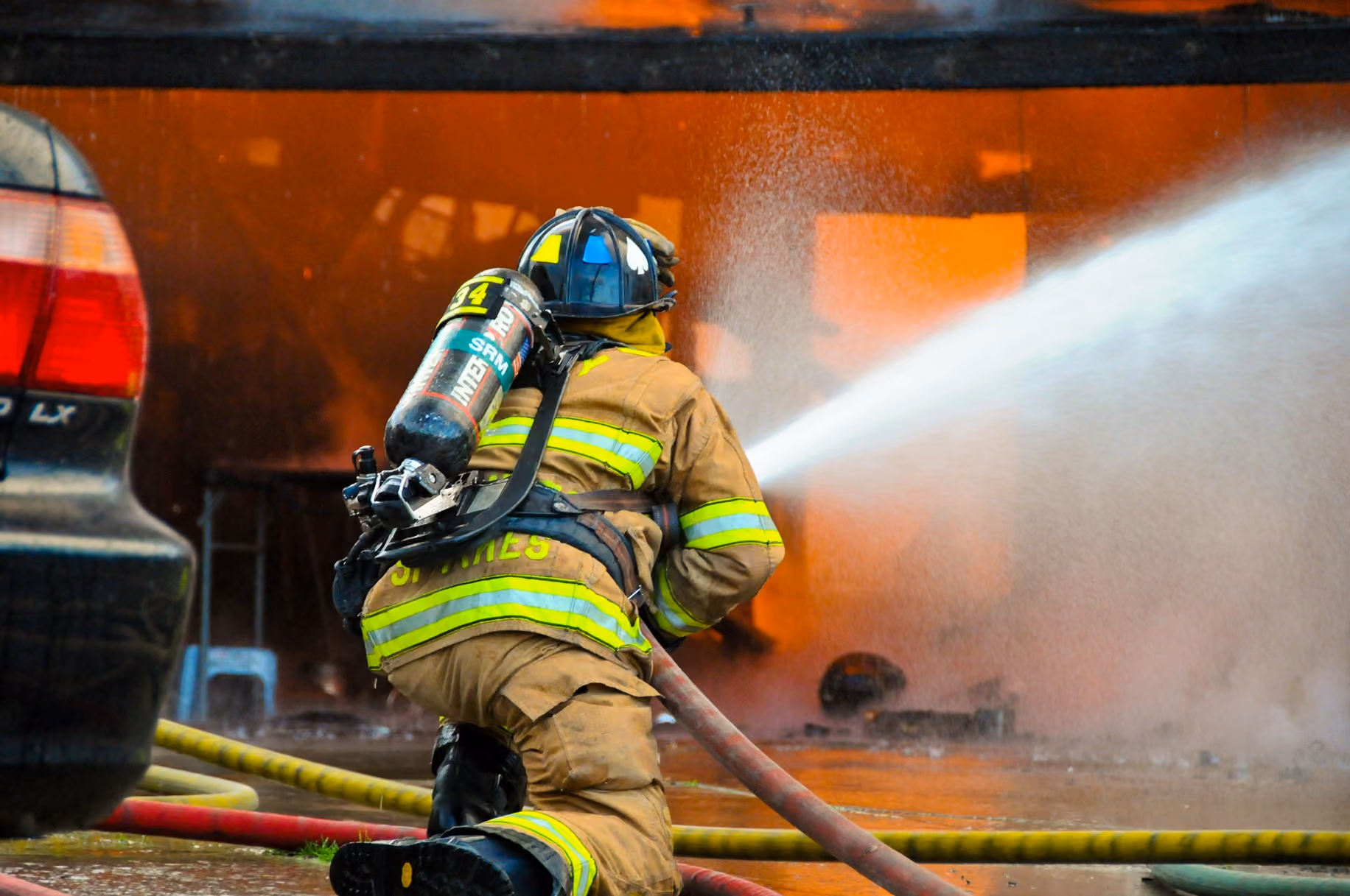 Firefighter in full gear kneels spraying water at a large orange fire through thick smoke.