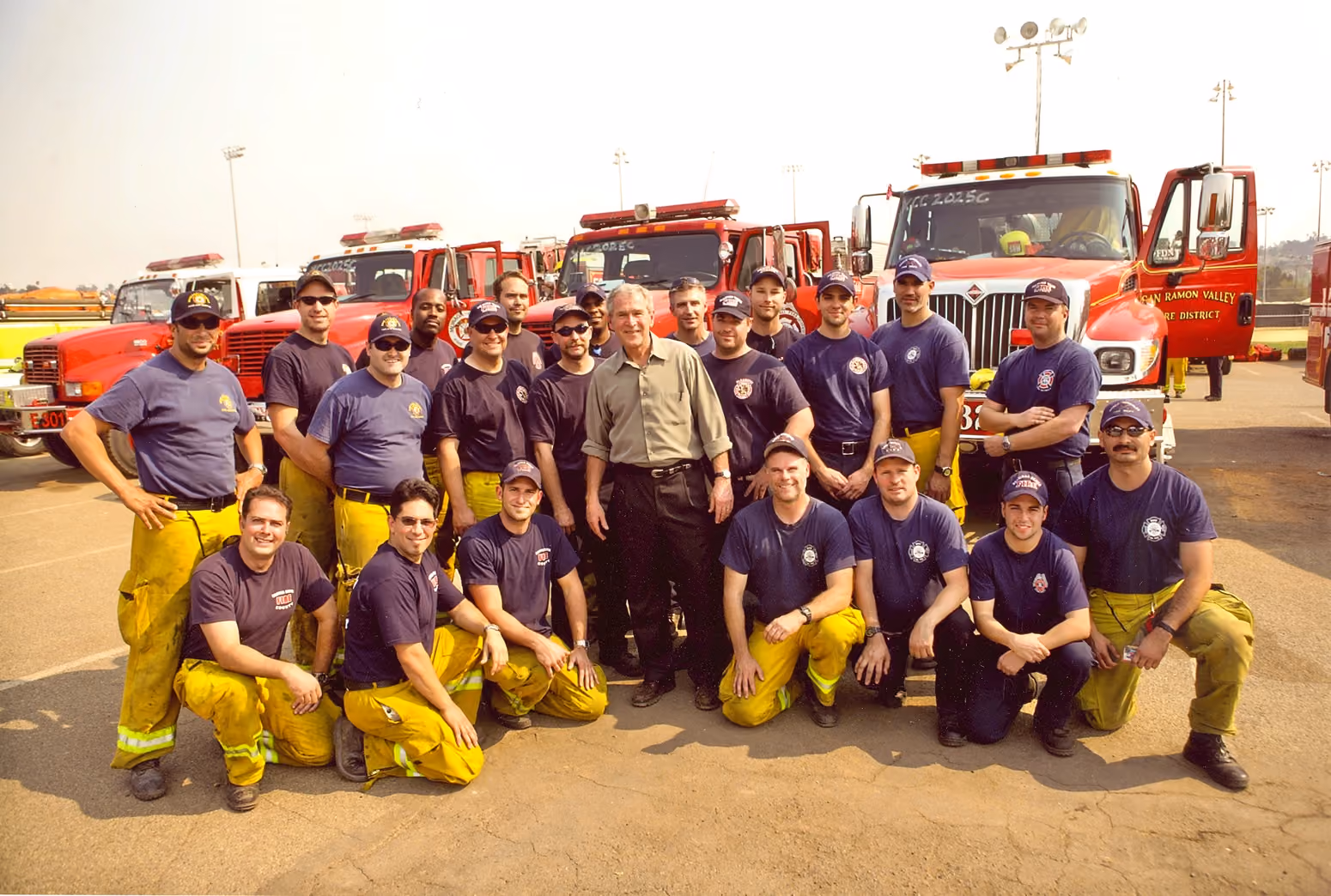 Group of firefighters in yellow pants and blue shirts posing with a man in tan shirt in front of fire trucks.