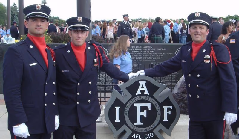 Three uniformed firefighters in dress uniforms posing by an International Association of Fire Fighters memorial plaque.