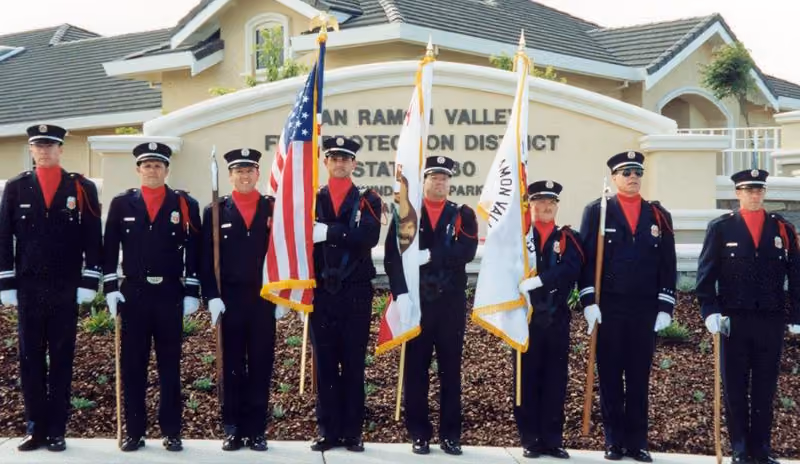 Seven uniformed honor guard members stand in a line holding the American and California flags.