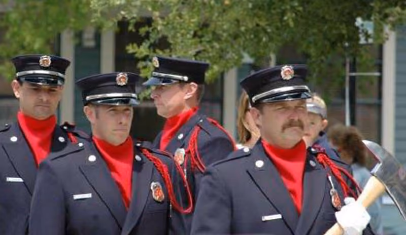 Four firefighters in dress uniforms with red turtlenecks, one holding an axe, at an outdoor event.