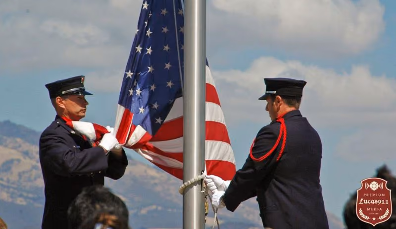 Two uniformed men folding an American flag beside a flagpole with a cloudy sky backdrop.