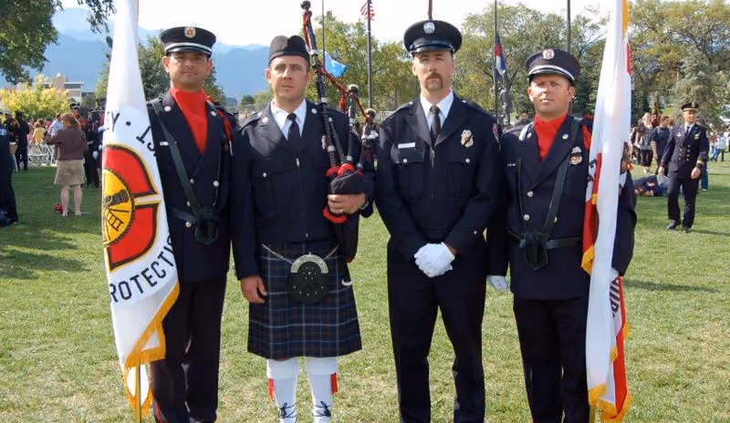 Four uniformed men standing on grass holding flags, one man wears a kilt and plays bagpipes.