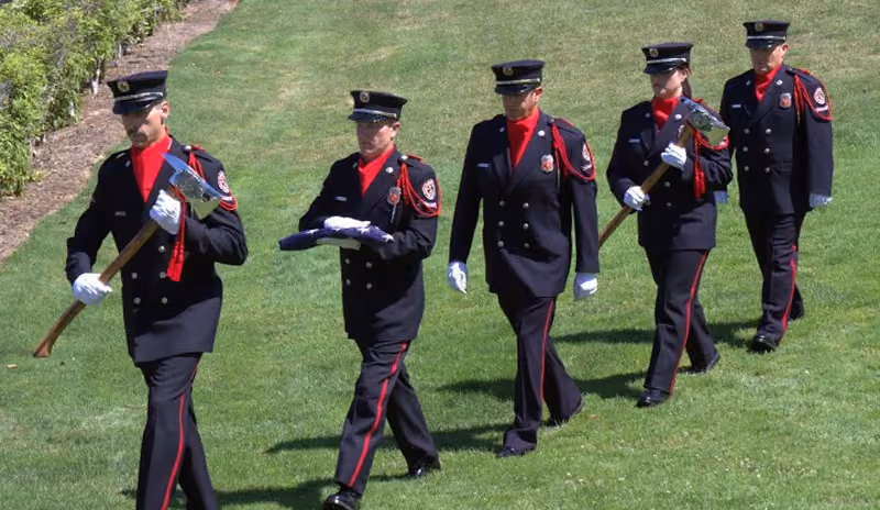 Five firefighters in formal dress uniforms march on grass, two holding ceremonial axes and one carrying folded flag.