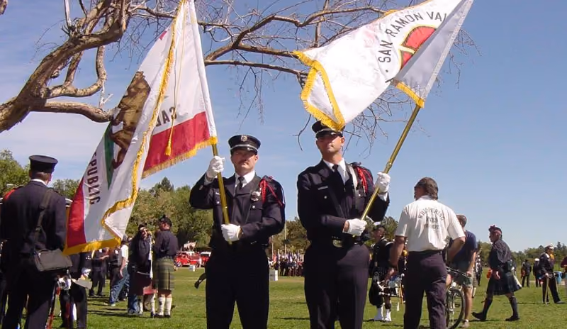 Two uniformed honor guard members holding flags during an outdoor ceremony on a sunny day.