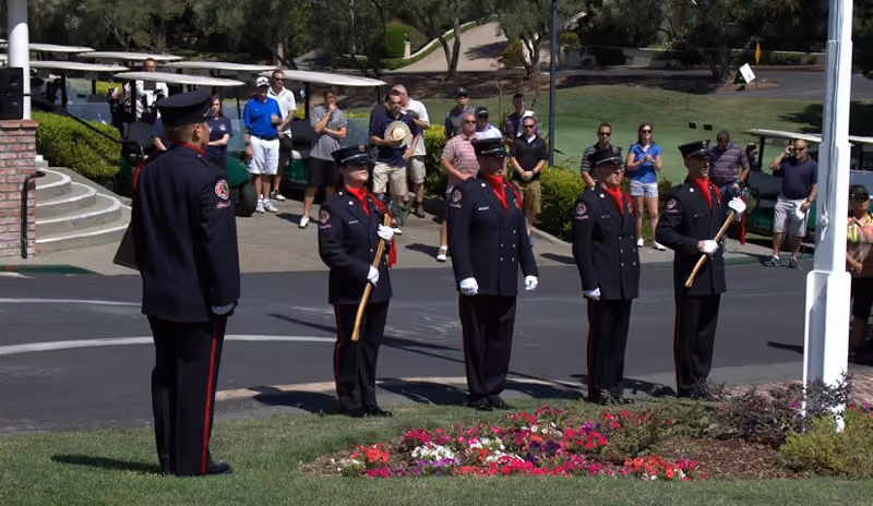 Honor guard in uniform standing solemnly near a flagpole with onlookers in the background.