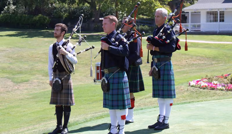 Four men wearing kilts and playing bagpipes outdoors on a grassy area near a building.