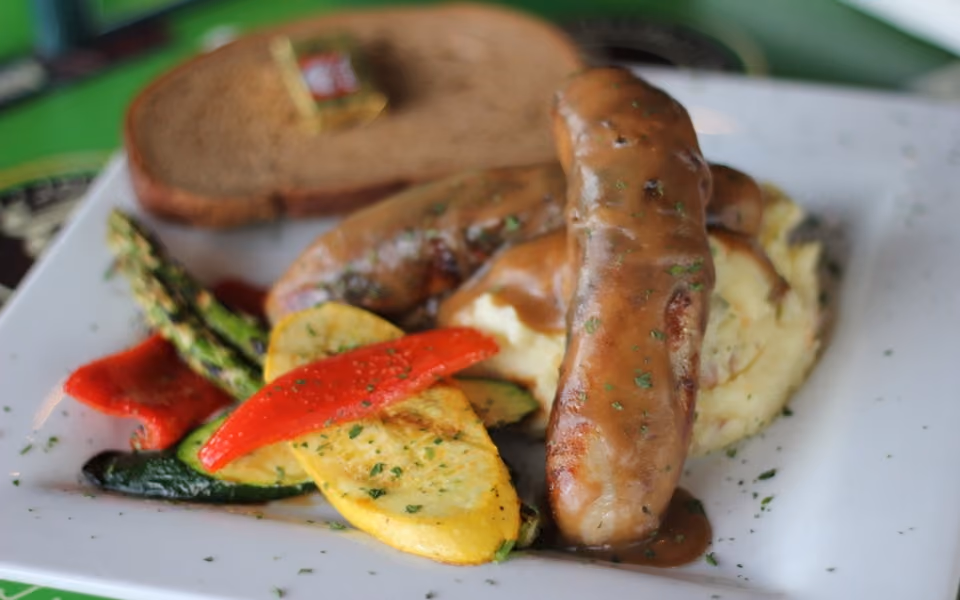 Plate with sausages and brown gravy over mashed potatoes, with grilled vegetables and a slice of bread.