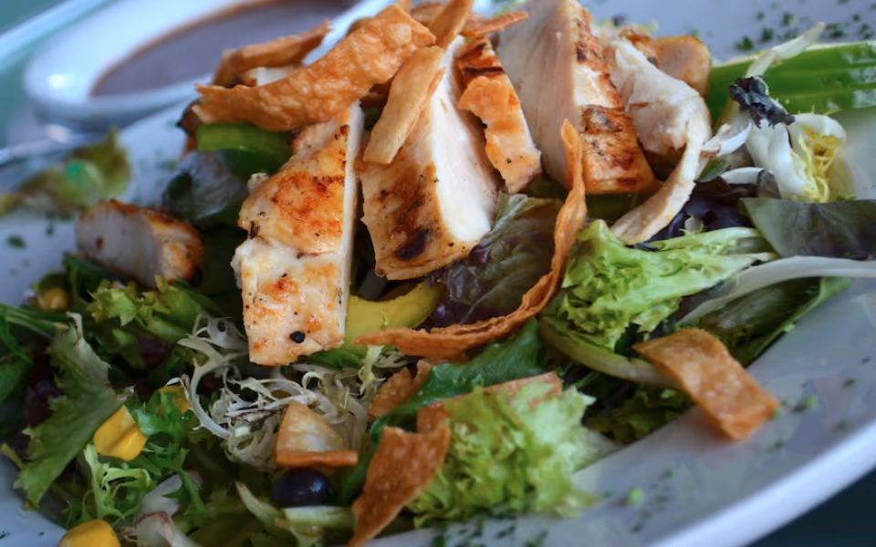 Grilled chicken salad with mixed greens, avocado, corn, and crispy tortilla strips on a white plate.