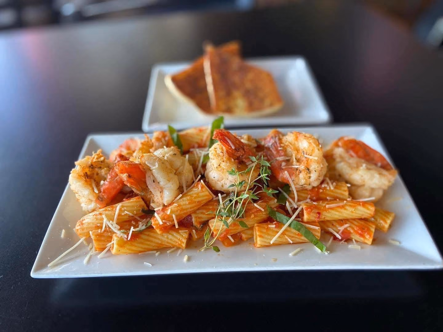 Plate of rigatoni pasta with shrimp, tomato sauce, herbs, and grated cheese with bread in the background.