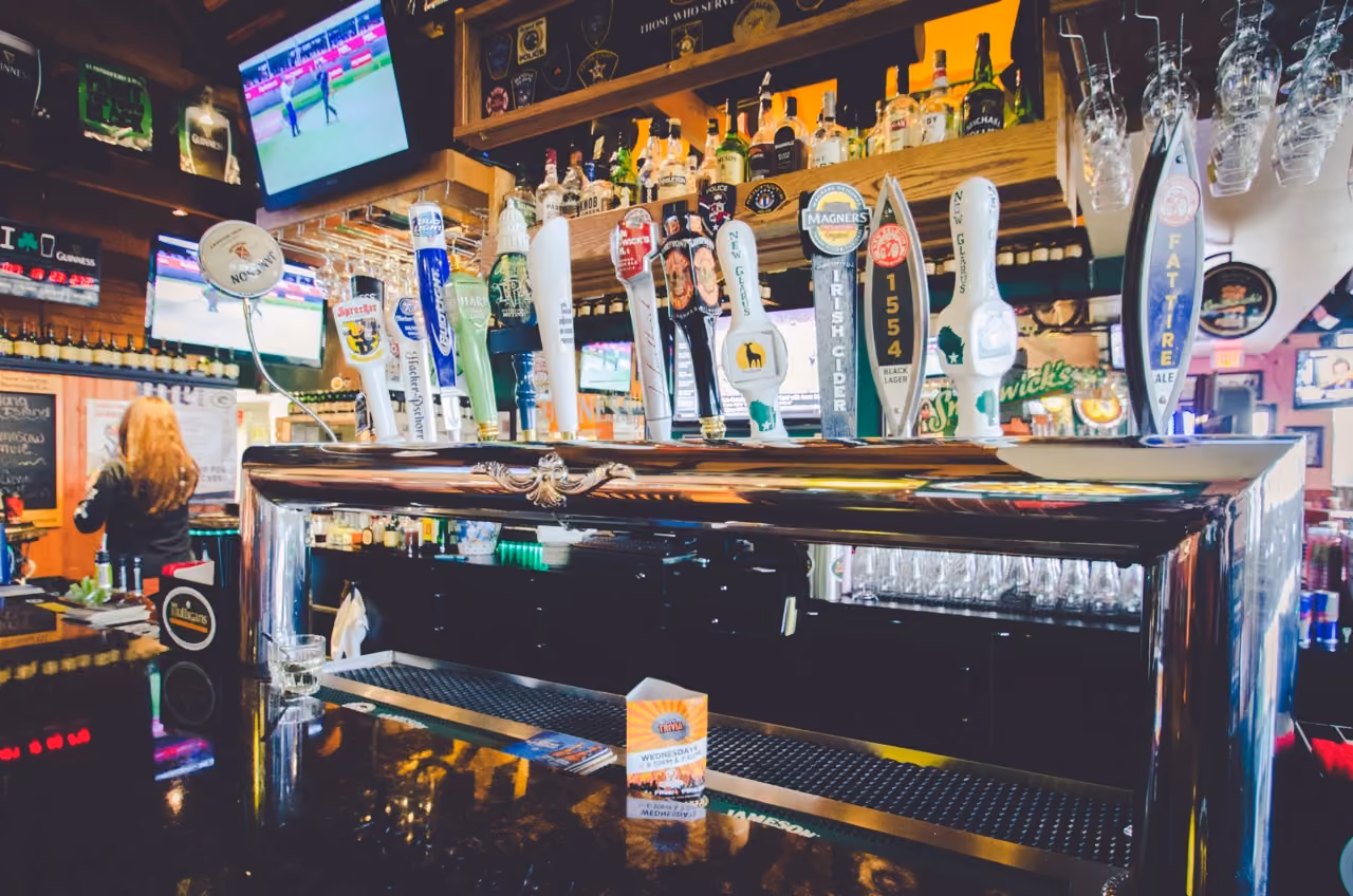 Variety of beer taps on shiny metal bar counter in an Irish pub with TV screens in the background.