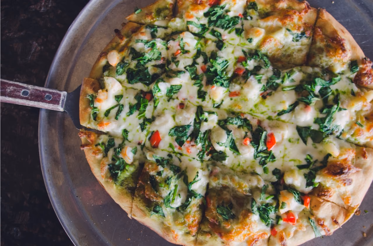 Pizza on metal tray with melted cheese, spinach, pesto sauce, and red pepper pieces sliced into squares.