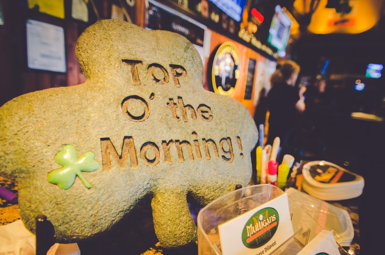 Stone shamrock sign on bar counter reading 'TOP O' the Morning!' with pub items and blurred background.