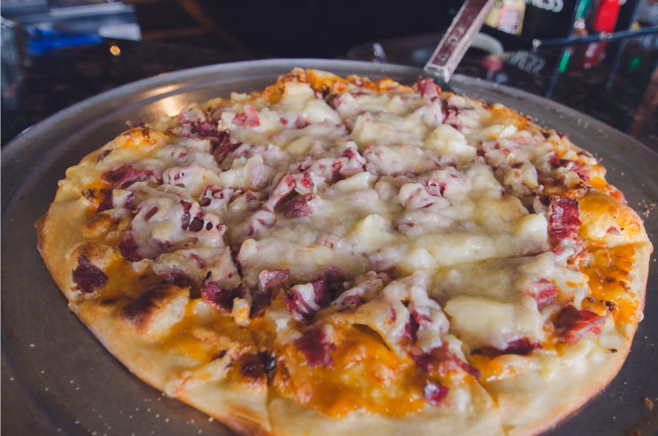 Close-up of a cheese pizza with bits of meat on a metal tray on a dark surface.