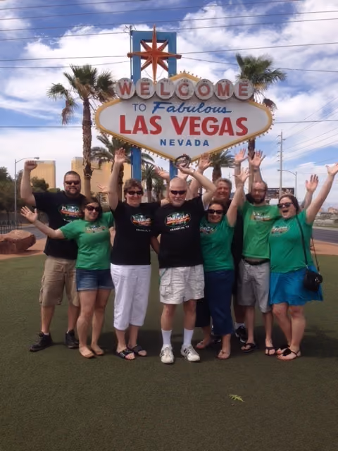 Group of eight people in matching shirts with arms raised in front of Welcome to Fabulous Las Vegas sign.