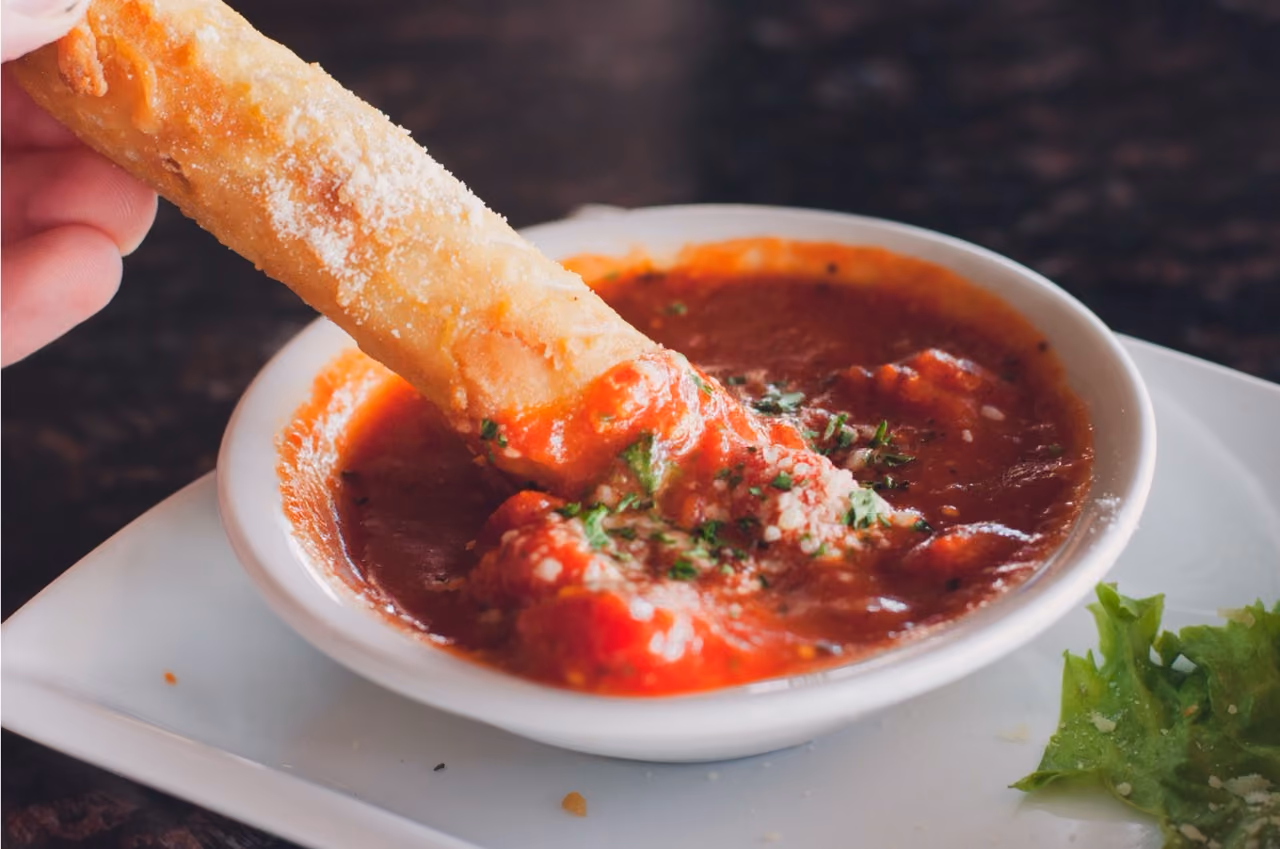 Hand dipping a fried breadstick into a bowl of marinara sauce topped with parsley and cheese.