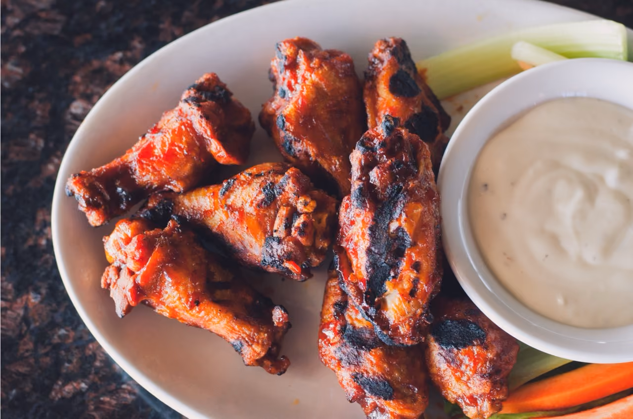 Grilled buffalo chicken wings with celery, carrot sticks, and creamy dipping sauce on a white plate.